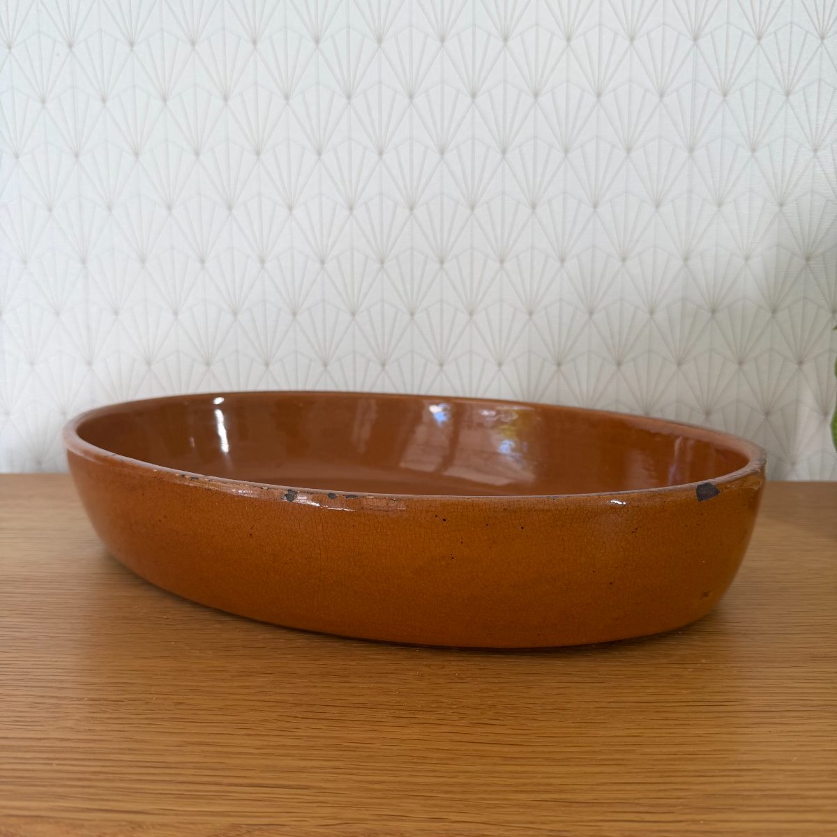Vintage French oval terracotta glazed baking dish in amber glaze, side profile view on oak surface against art deco wallpaper.
