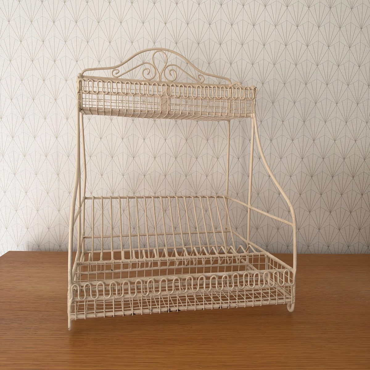 Vintage French white painted wire metal two-tier dish rack egouttoir with ornate scrollwork crest, shown on a wood table against art deco wallpaper.