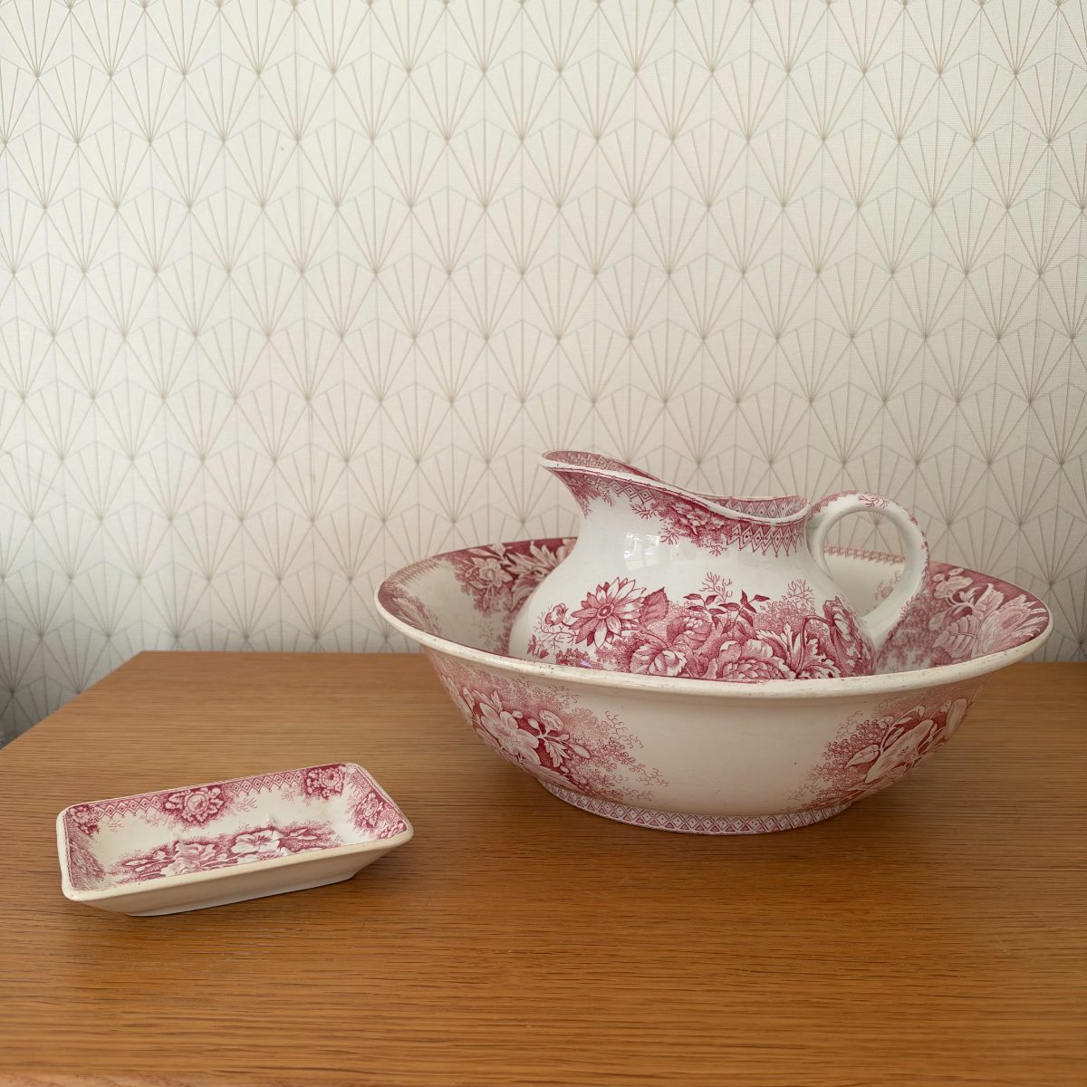 Full view of French red transferware pitcher and wash bowl set with soap dish on wooden surface against art deco wallpaper.