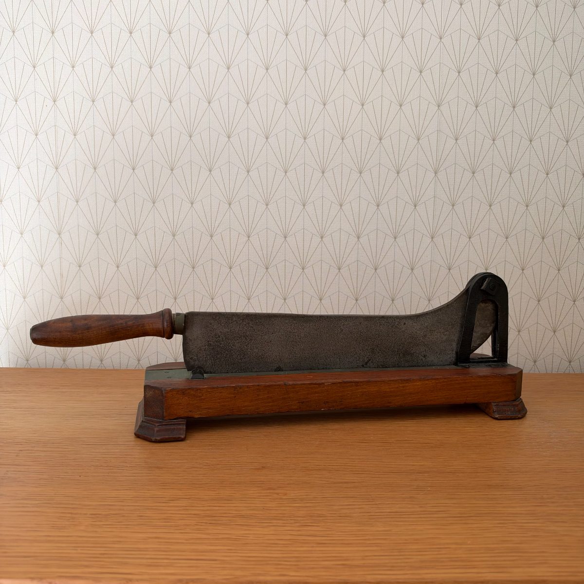 Antique French cast iron bread slicer on dark walnut wooden base with turned wood handle, side profile view on oak table.