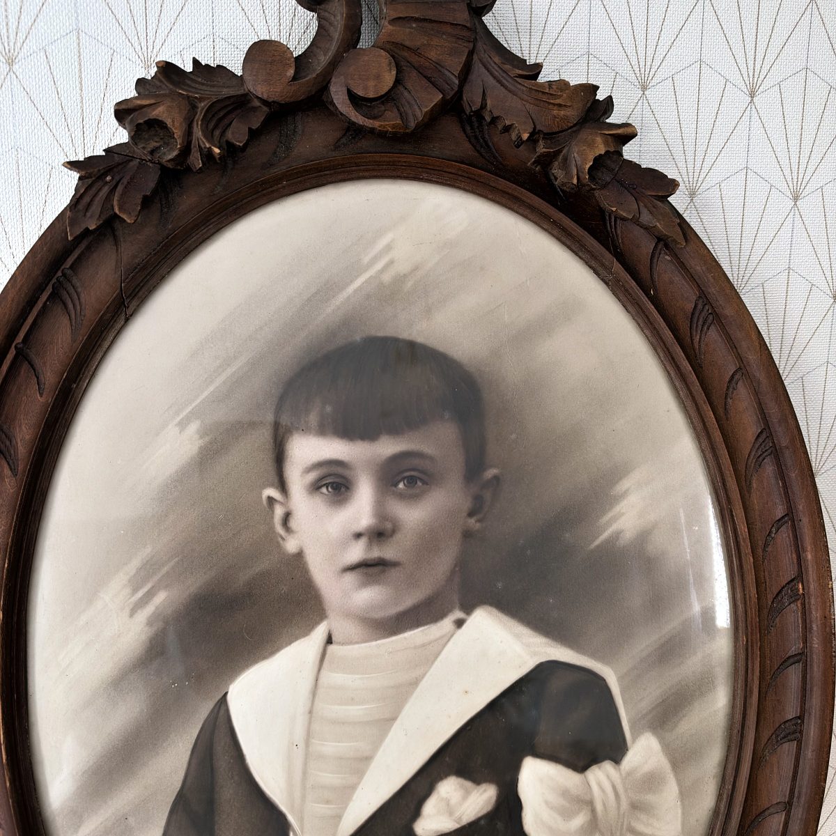 Close-up of antique French carved wood oval frame showing boy in sailor suit with bow, convex glass over sepia portrait, ornate crest detail at top.