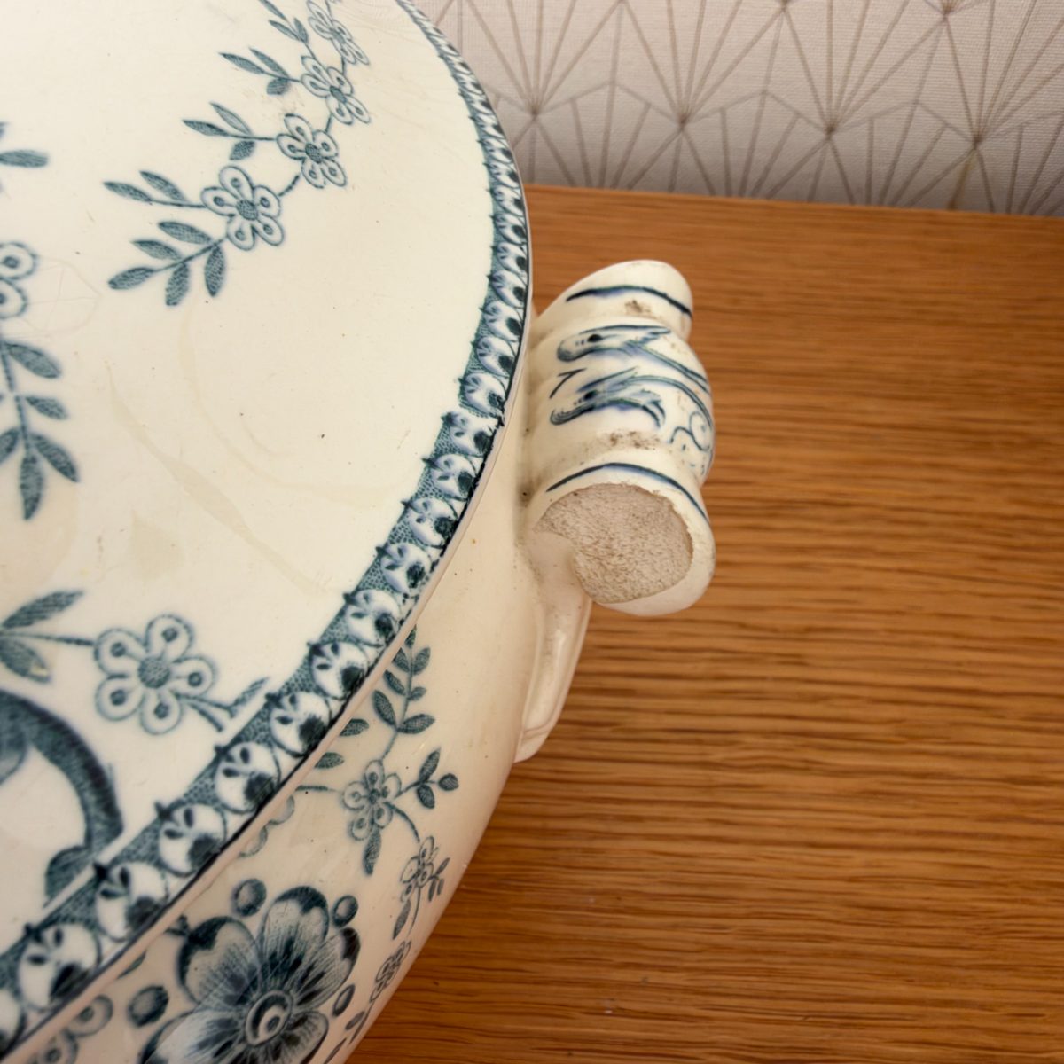Top-down close-up of chipped handle on antique French blue white faience soup tureen, showing blue scrollwork decoration and exposed clay.
