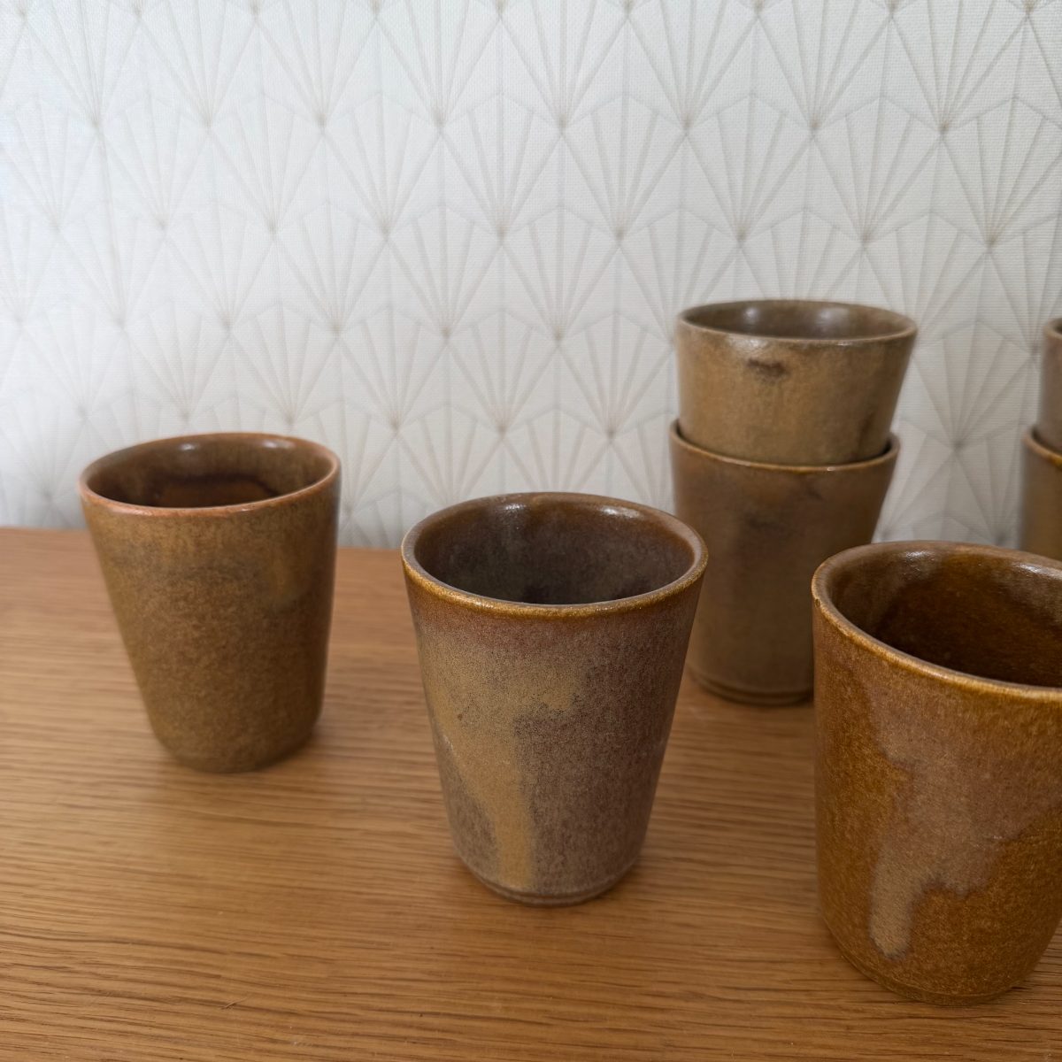 Close-up of three French studio stoneware cups with matte amber glaze and dark interior, two stacked in background on oak table.