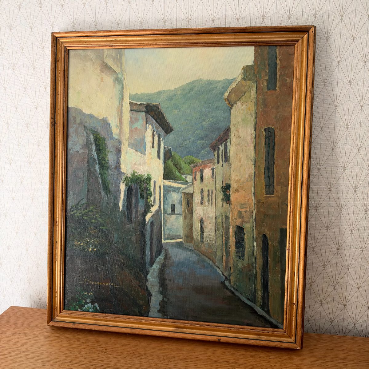 Full front view of framed French oil painting by Dorgendial depicting a curved village street with stone facades and mountain backdrop.