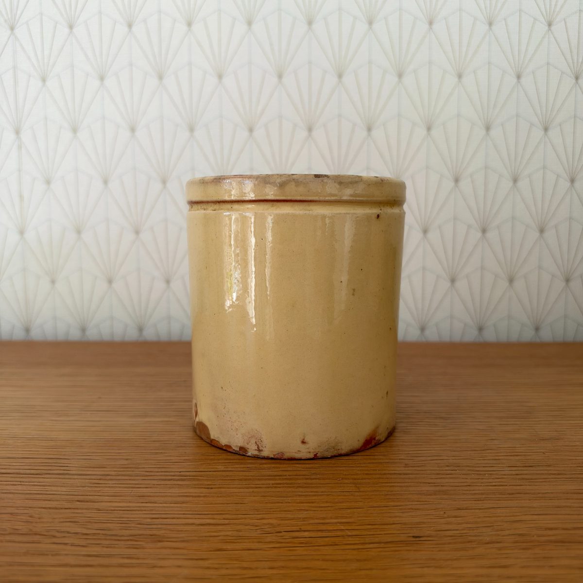 Front view of antique French stoneware jam pot with golden yellow glaze and raised rim, sitting on a wood surface against Art Deco wallpaper.
