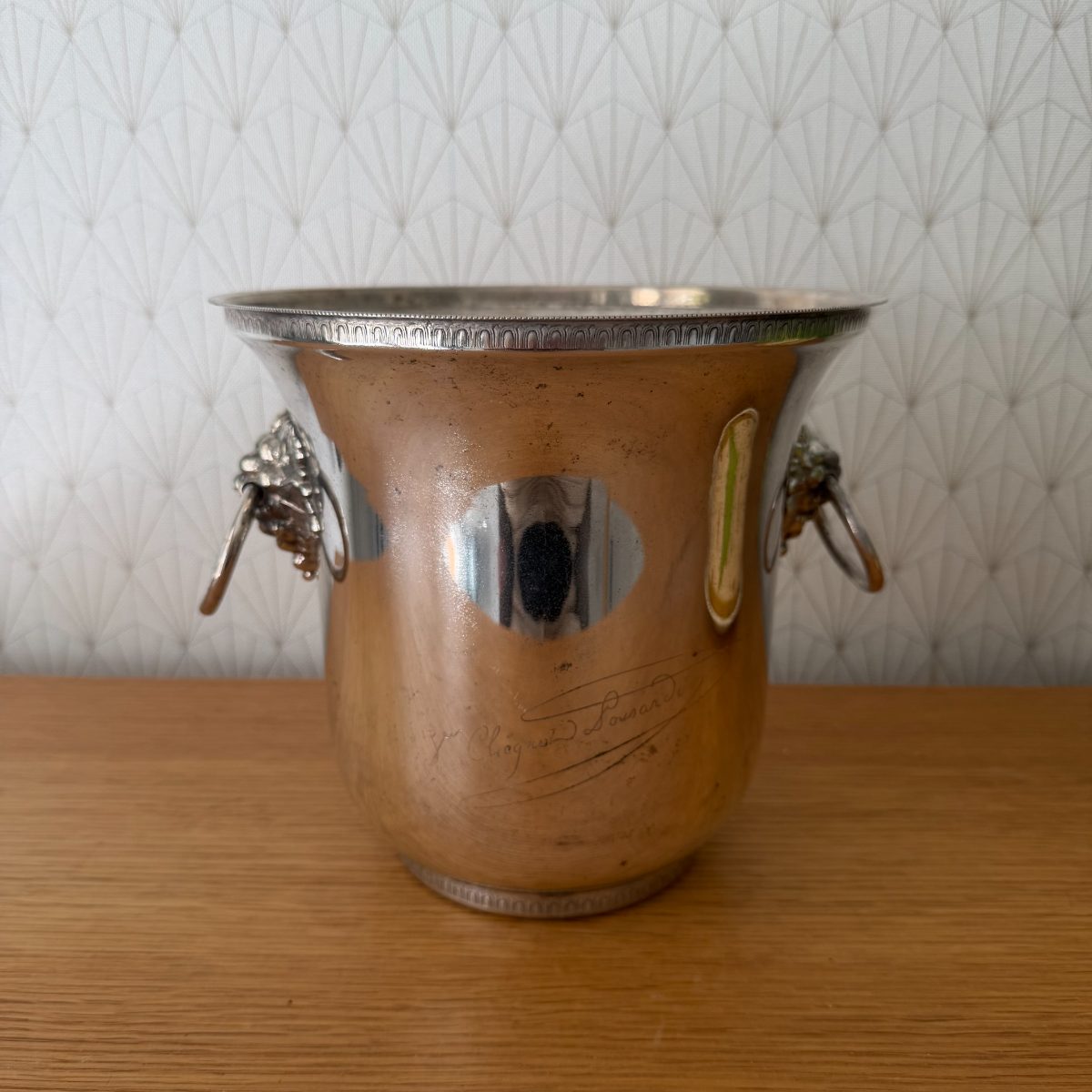 French silver plated Veuve Clicquot champagne bucket showing tarnished patina and lion ring handles, side angle on wooden table.