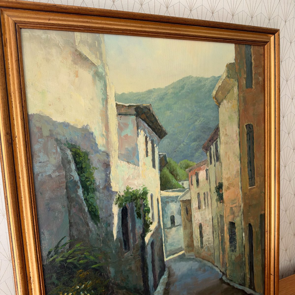 Close-up upper portion of oil painting showing sunlit whitewashed buildings, green hills backdrop and ivy-covered stone wall, French village scene.