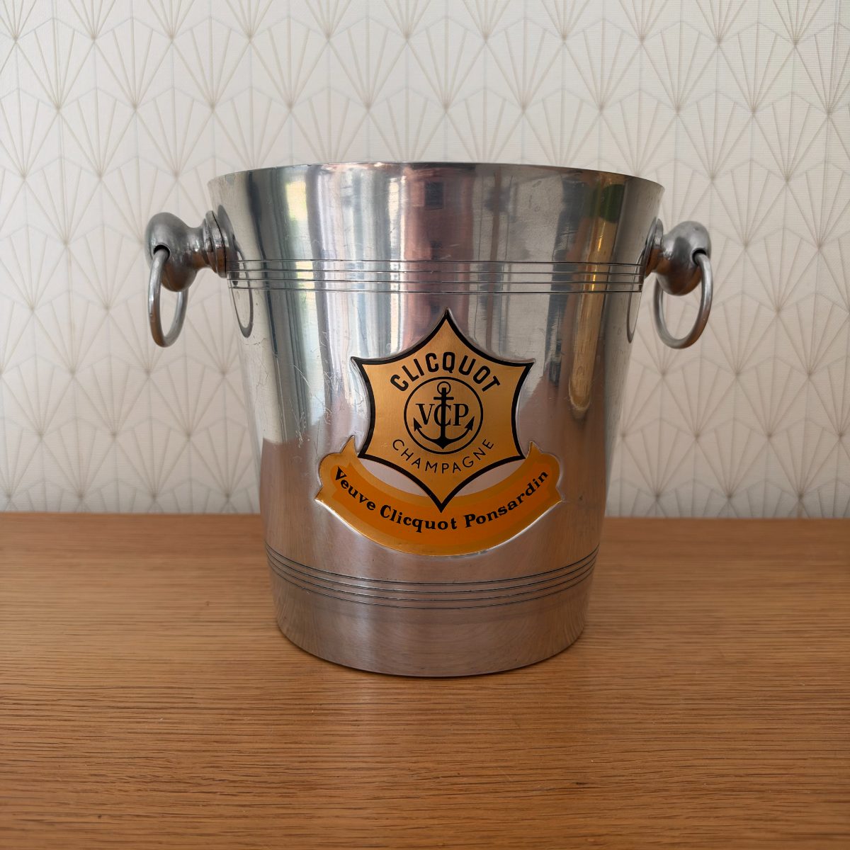 Front view of vintage French Veuve Clicquot Ponsardin aluminium champagne ice bucket with orange VCP shield badge and ring handles on oak surface.