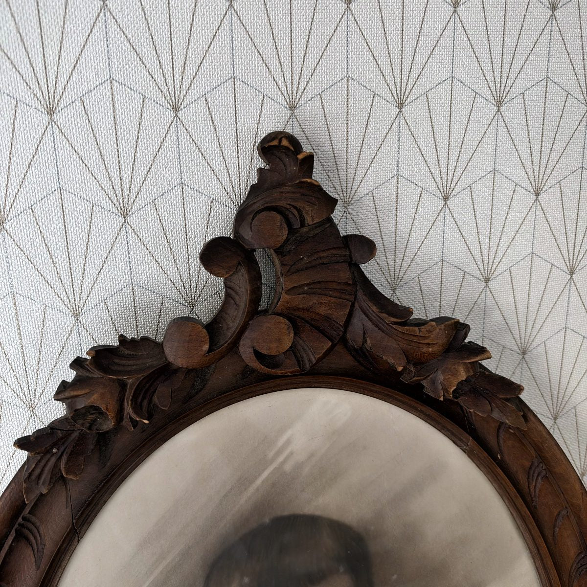 Detail of carved wooden crest at top of antique French oval frame, showing acanthus scrolls and leaf motifs in dark walnut-toned wood.