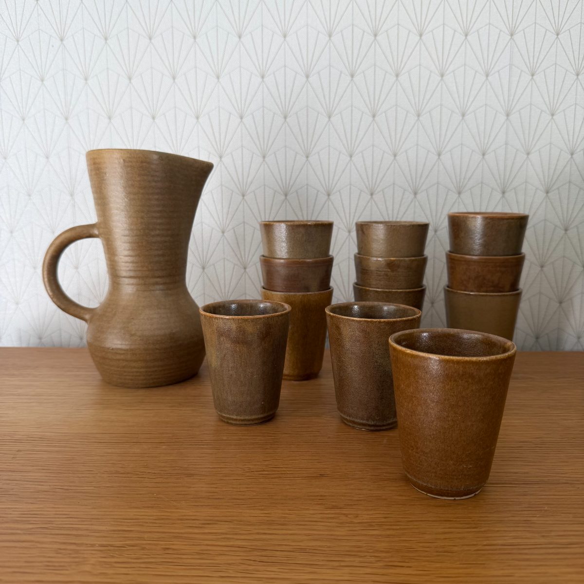 Vintage French stoneware pitcher and twelve cups set in ochre and amber glaze displayed on a wood surface against geometric wallpaper