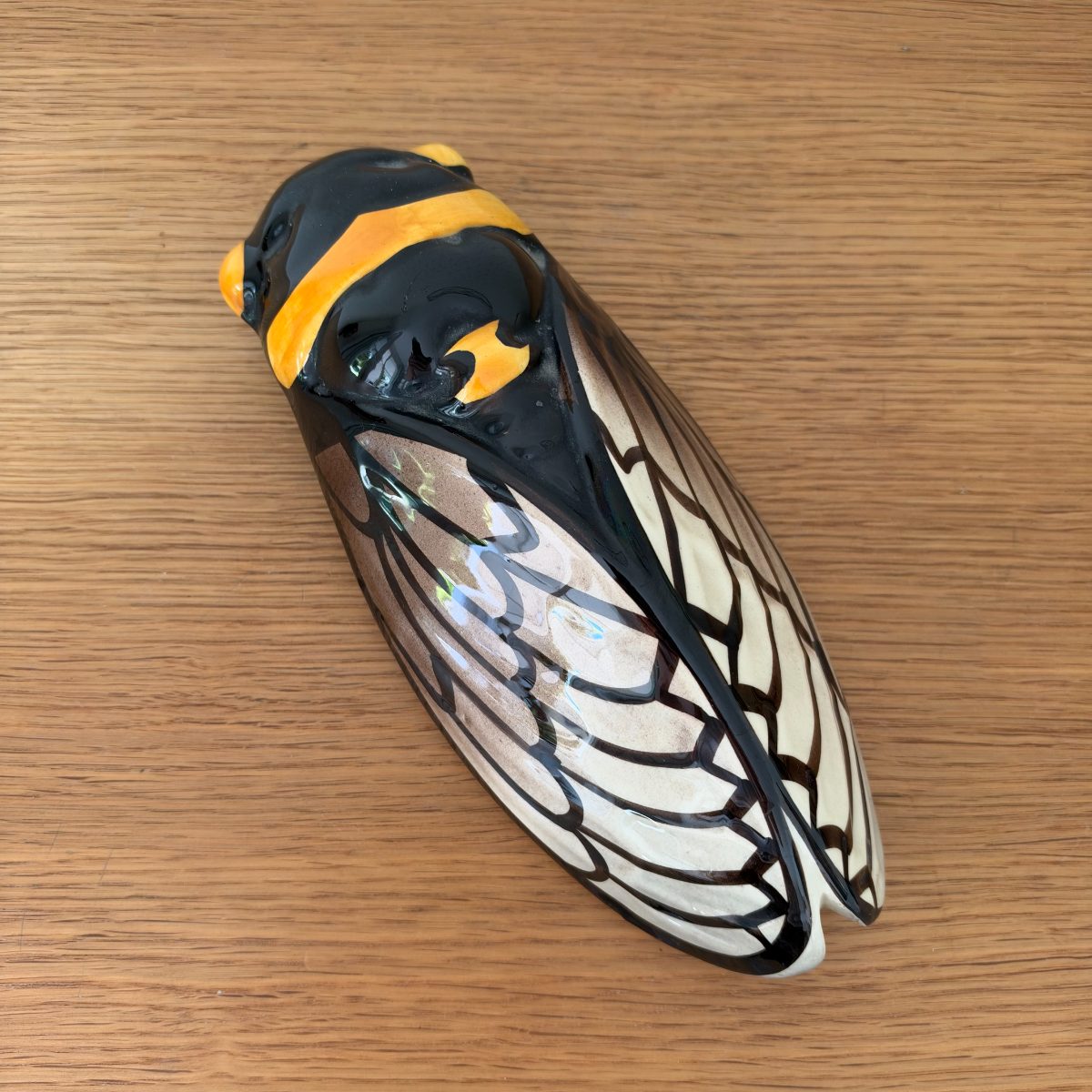 Top-down view of French ceramic cicada figurine with veined wing glaze in cream and black, amber orange head markings, on oak table.