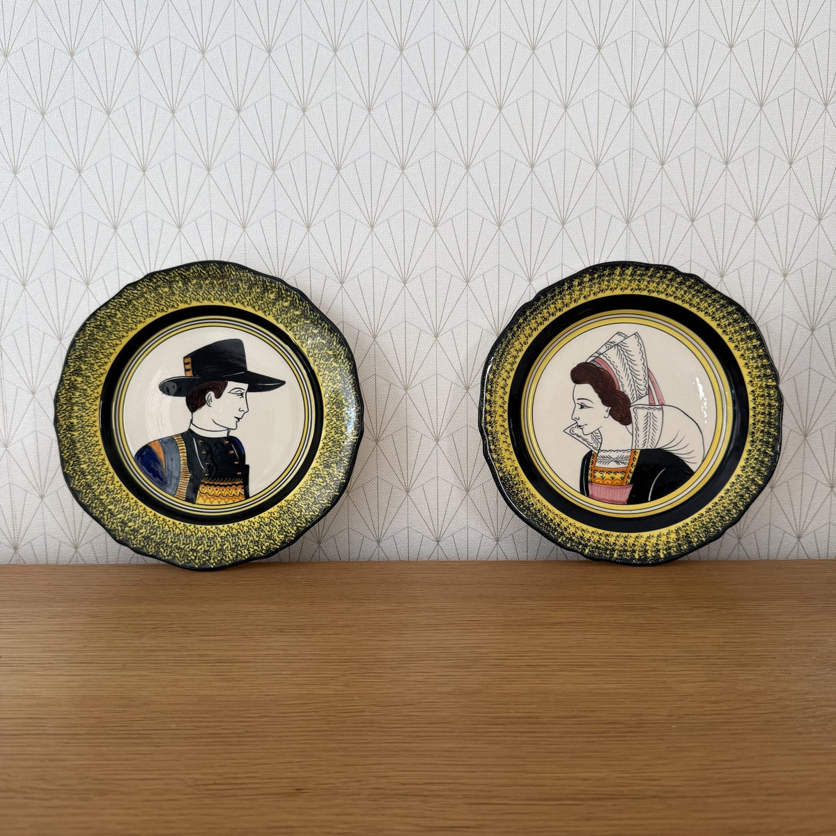 Pair of Henriot Quimper portrait plates with hand-painted Breton man in black hat and woman in traditional dress against patterned background