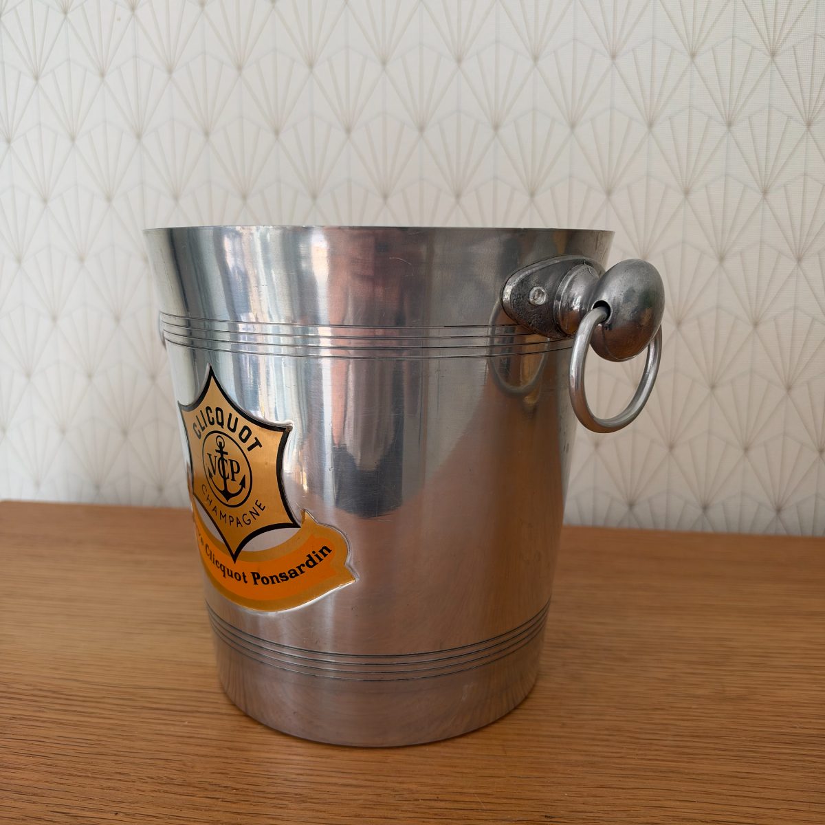 Side angle view of Veuve Clicquot Ponsardin aluminium champagne ice bucket showing ring handle and partial badge detail.