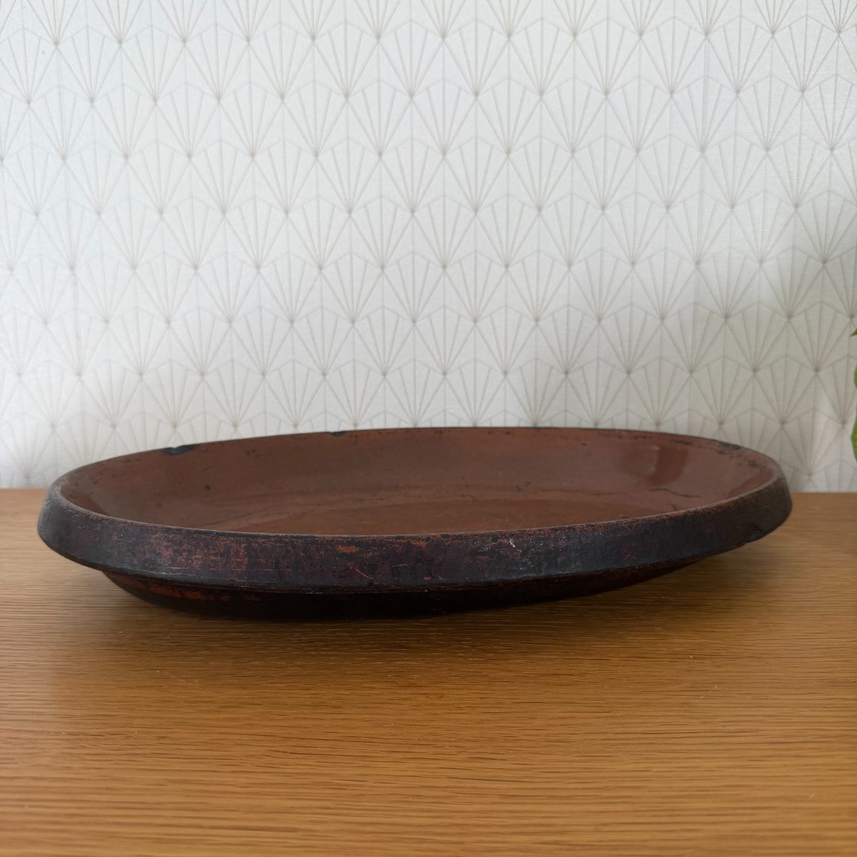 Side view of a vintage French oval glazed terracotta baking dish with dark rim and warm brown interior, placed on a wood surface.