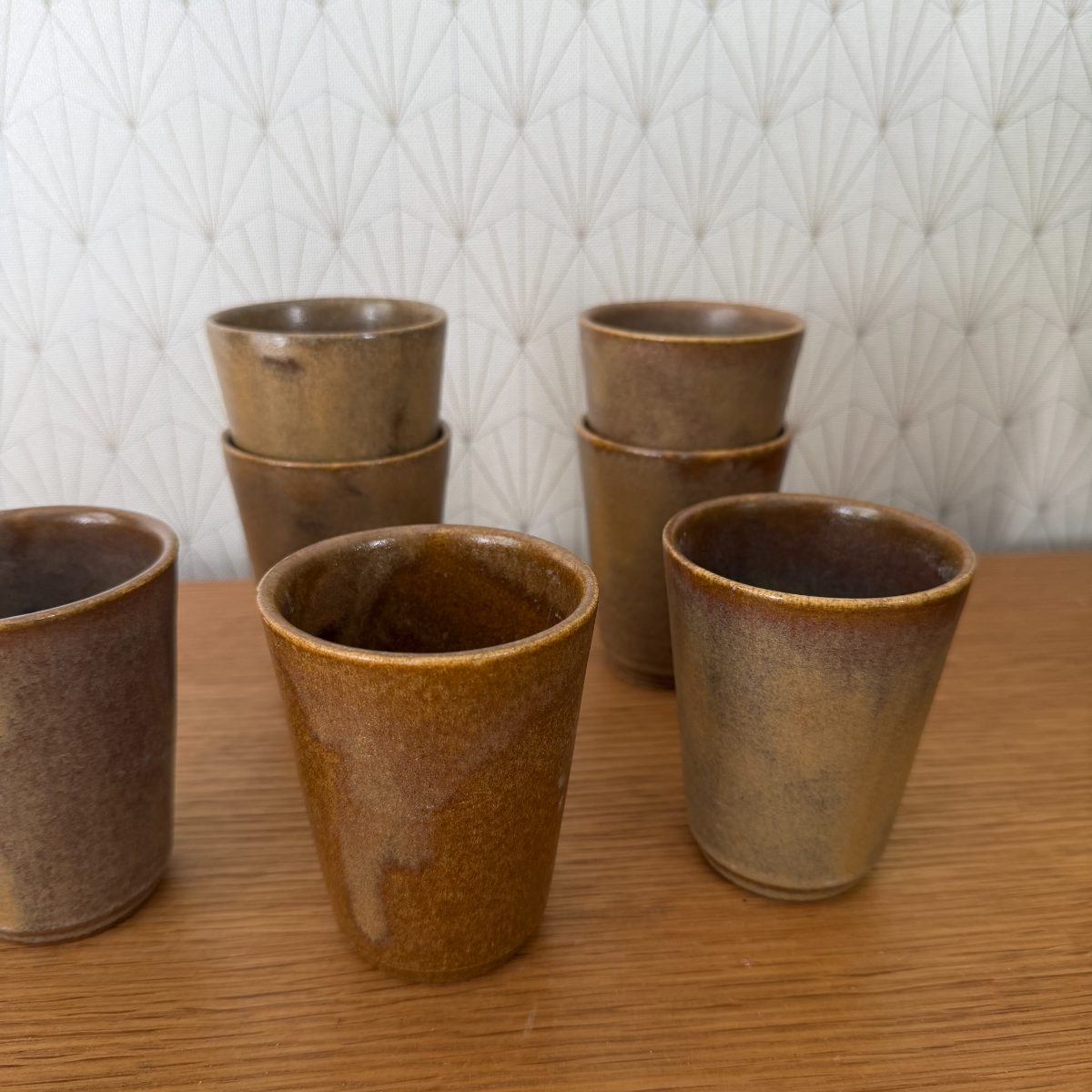 Eight French glazed stoneware cups arranged on a wood table, front row upright showing varied amber and ochre glaze tones.