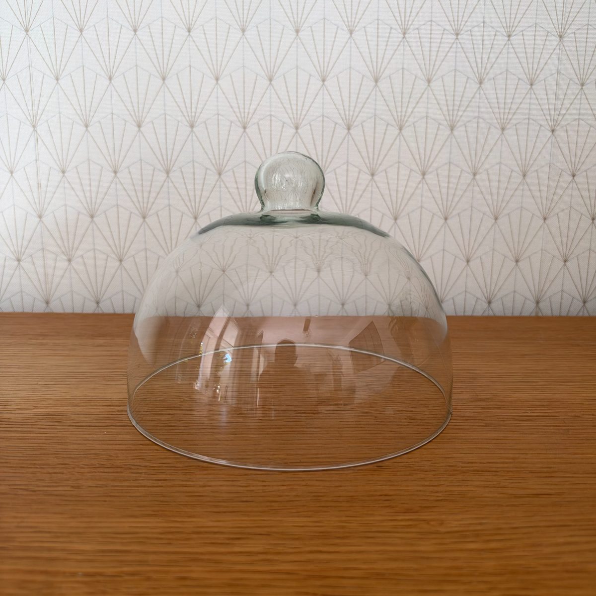 Vintage French clear glass cloche dome with round knob handle, sitting upright on a wood surface, art deco patterned wallpaper background, front view showing full profile.