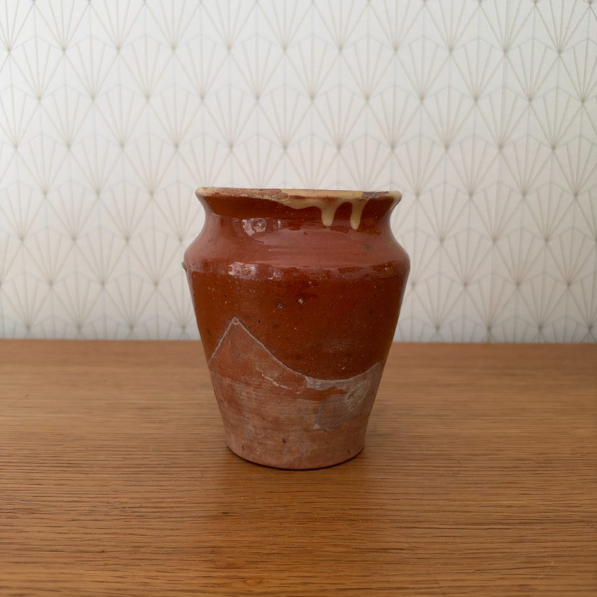 Front view of antique French earthenware pot with red-brown glaze over terracotta body, cream glaze drip at rim, partial glaze on lower half, on wood surface.