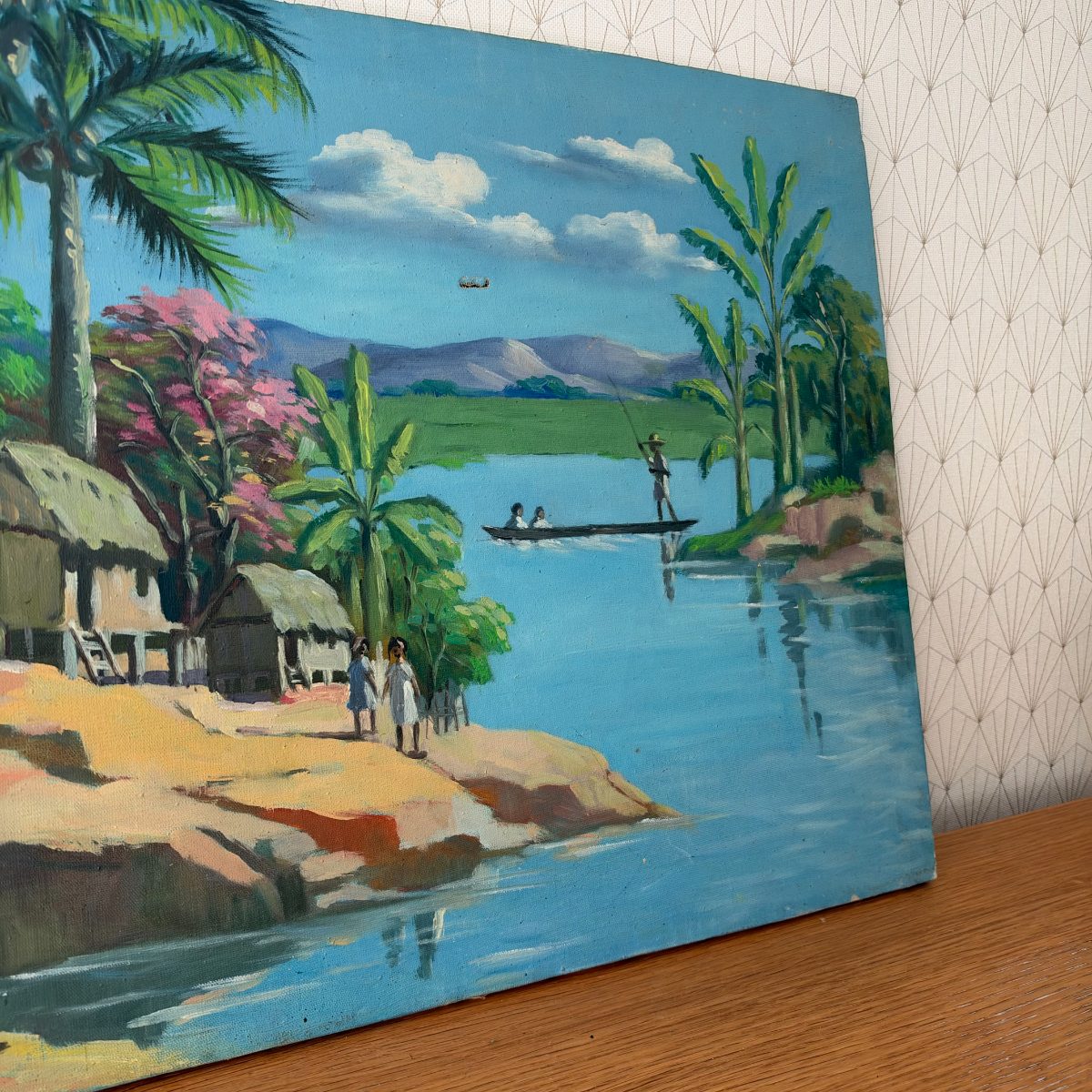 Right side view of vintage tropical oil painting on canvas showing river scene with fisherman on boat, mountains in background, and figures on the shore