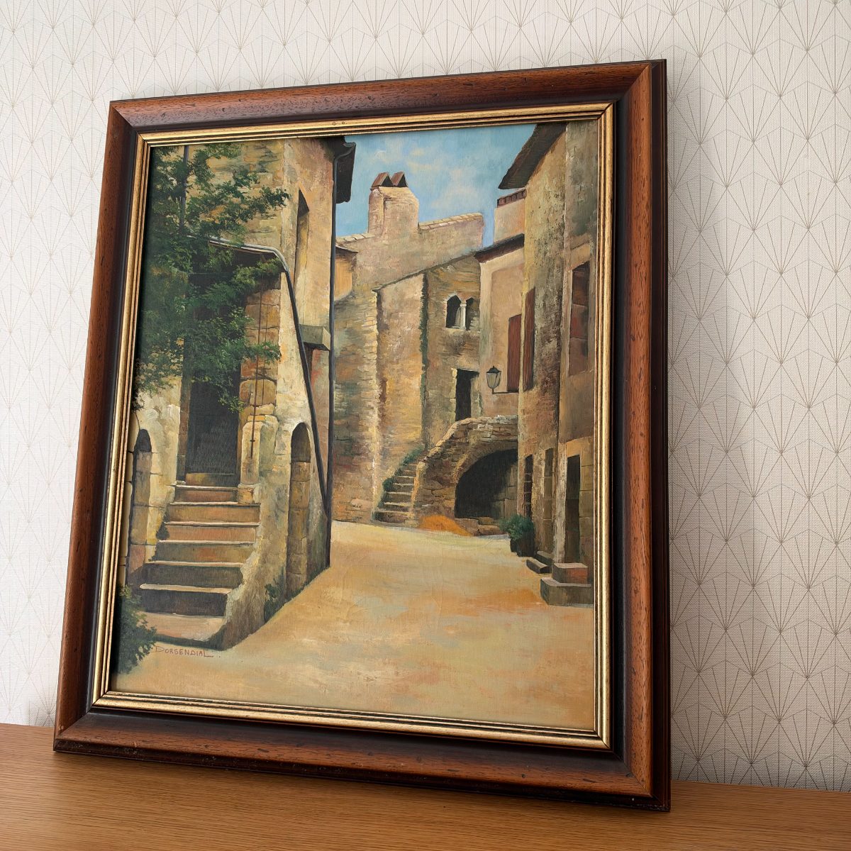 Side angle view of the complete framed French oil painting of a Gard village scene by Dorgendial on a wooden surface against patterned wallpaper.