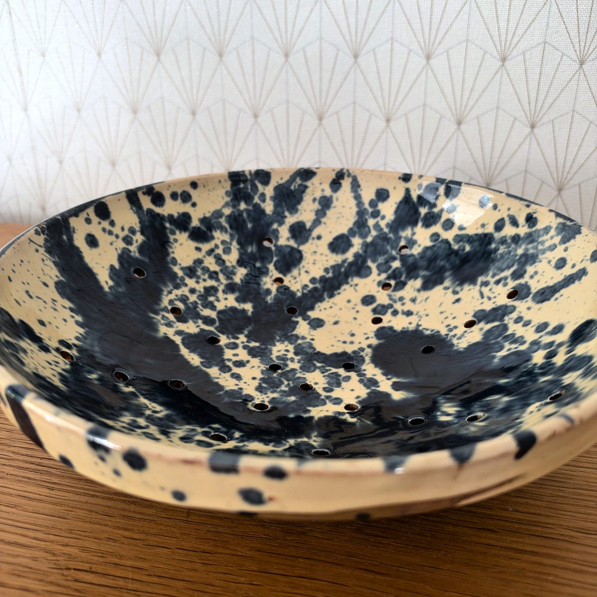 Angled interior view of French ceramic berry colander showing hand-pierced drainage holes and graphic black splatter glaze pattern on ochre ground.