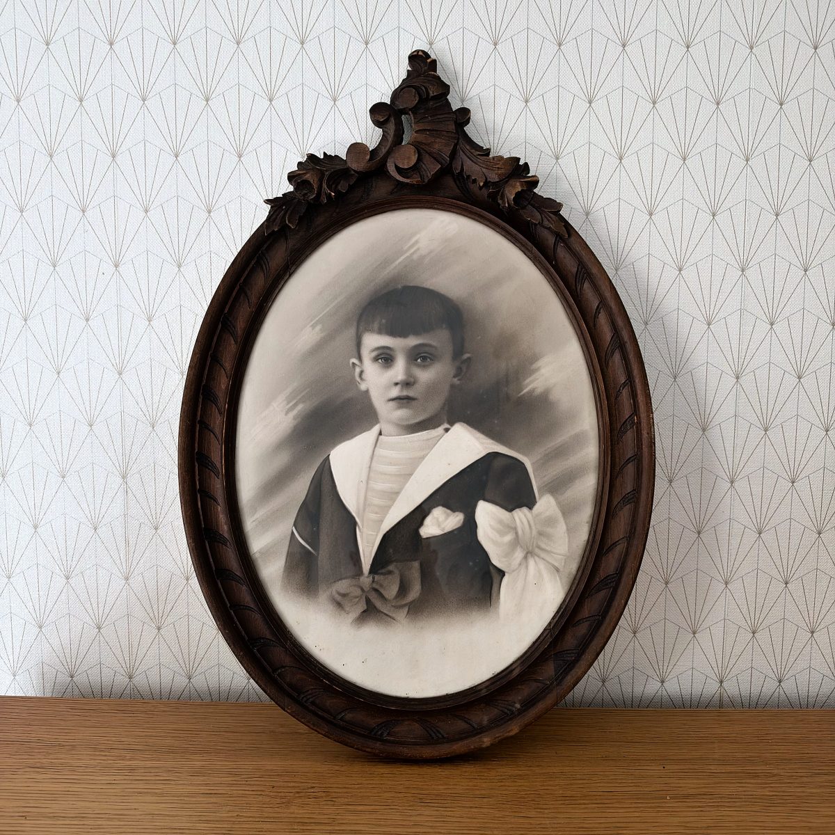 Antique French oval carved walnut frame with boy first communion portrait photo, decorative acanthus leaf crest, leaning on wooden shelf against geometric wallpaper.