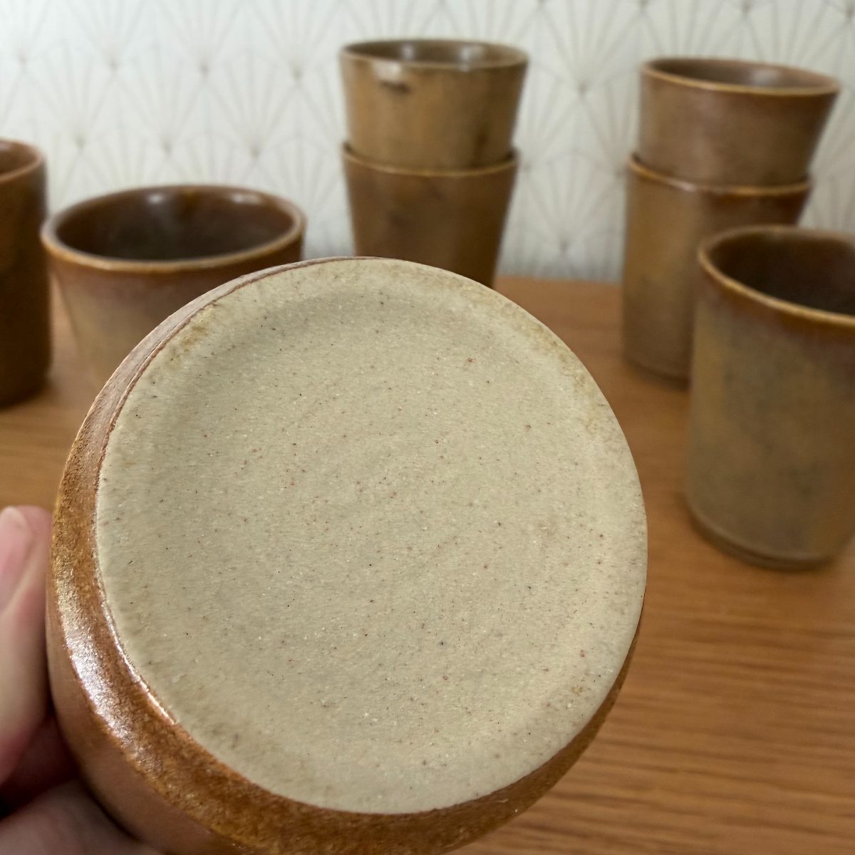 Unglazed foot of a French stoneware studio pottery cup held up showing speckled raw clay base, remaining cups visible in background.