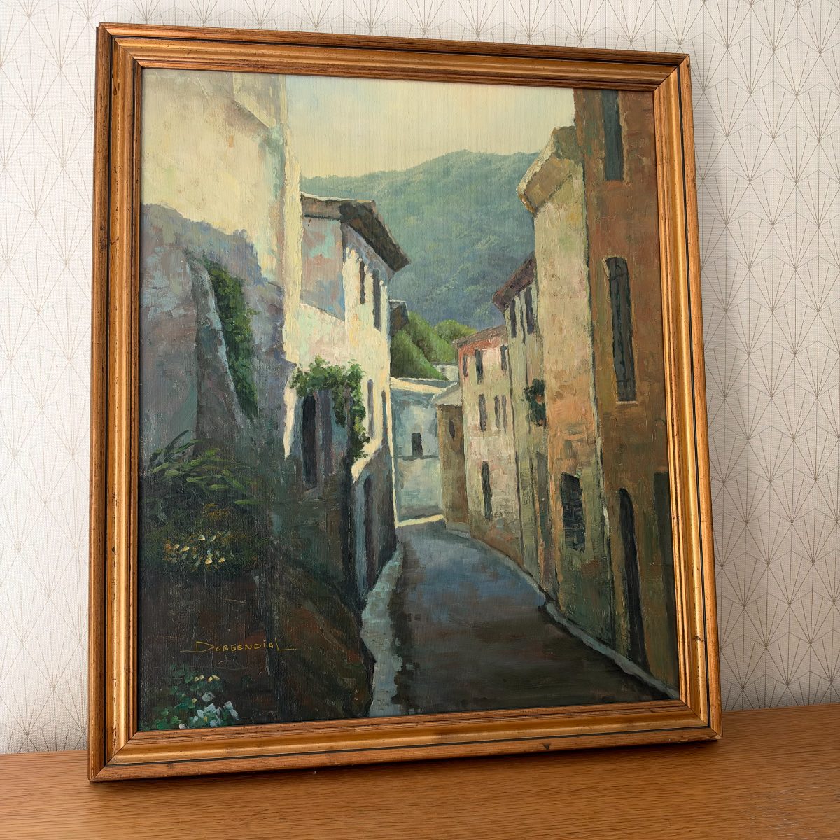Side angle view of framed French oil painting showing village alley with stone buildings and green hills, signed Dorgendial, gold frame.