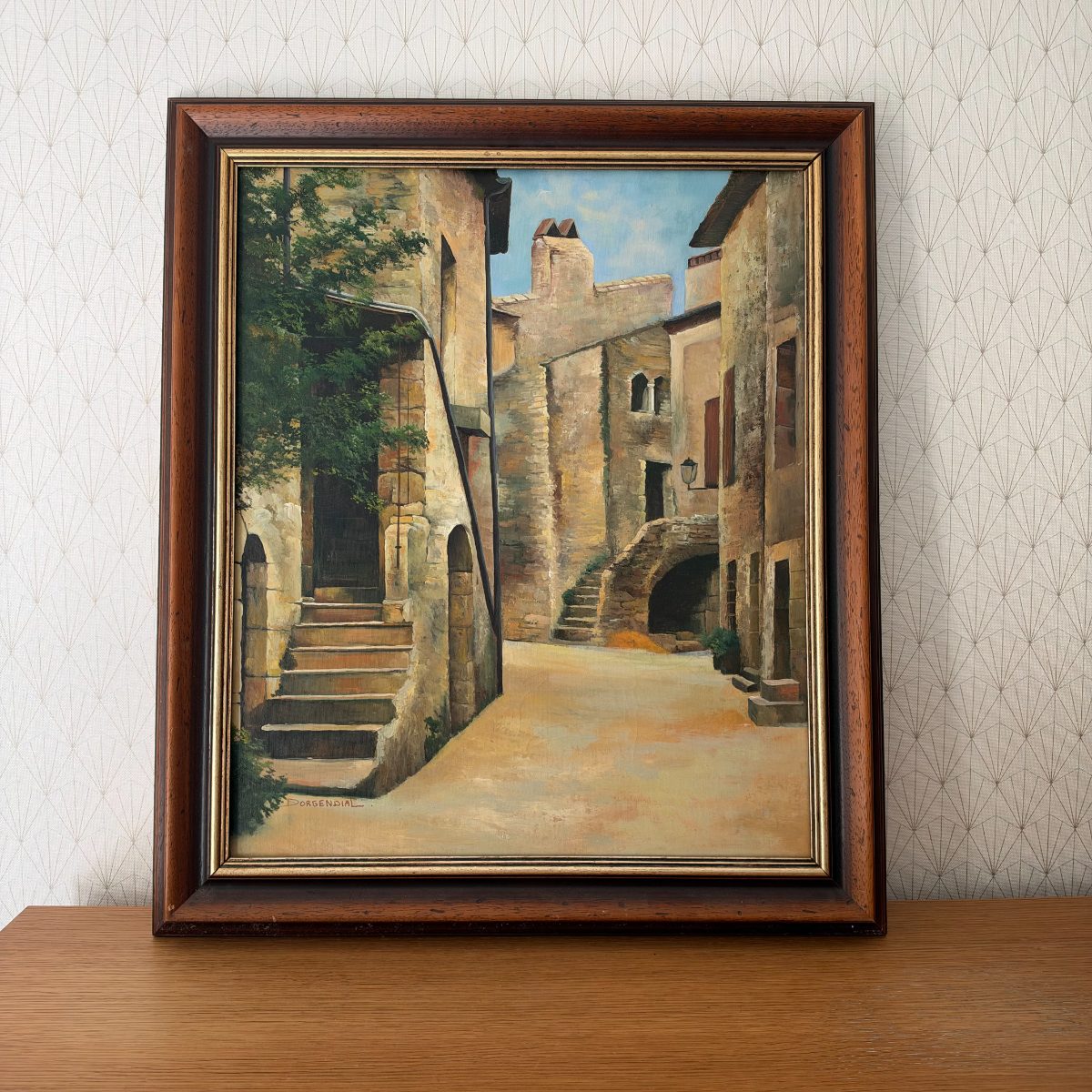 Framed French oil painting of a stone village street scene signed Dorgendial, warm ochre tones, dark wood frame with gold inner edge, leaning on wooden surface.