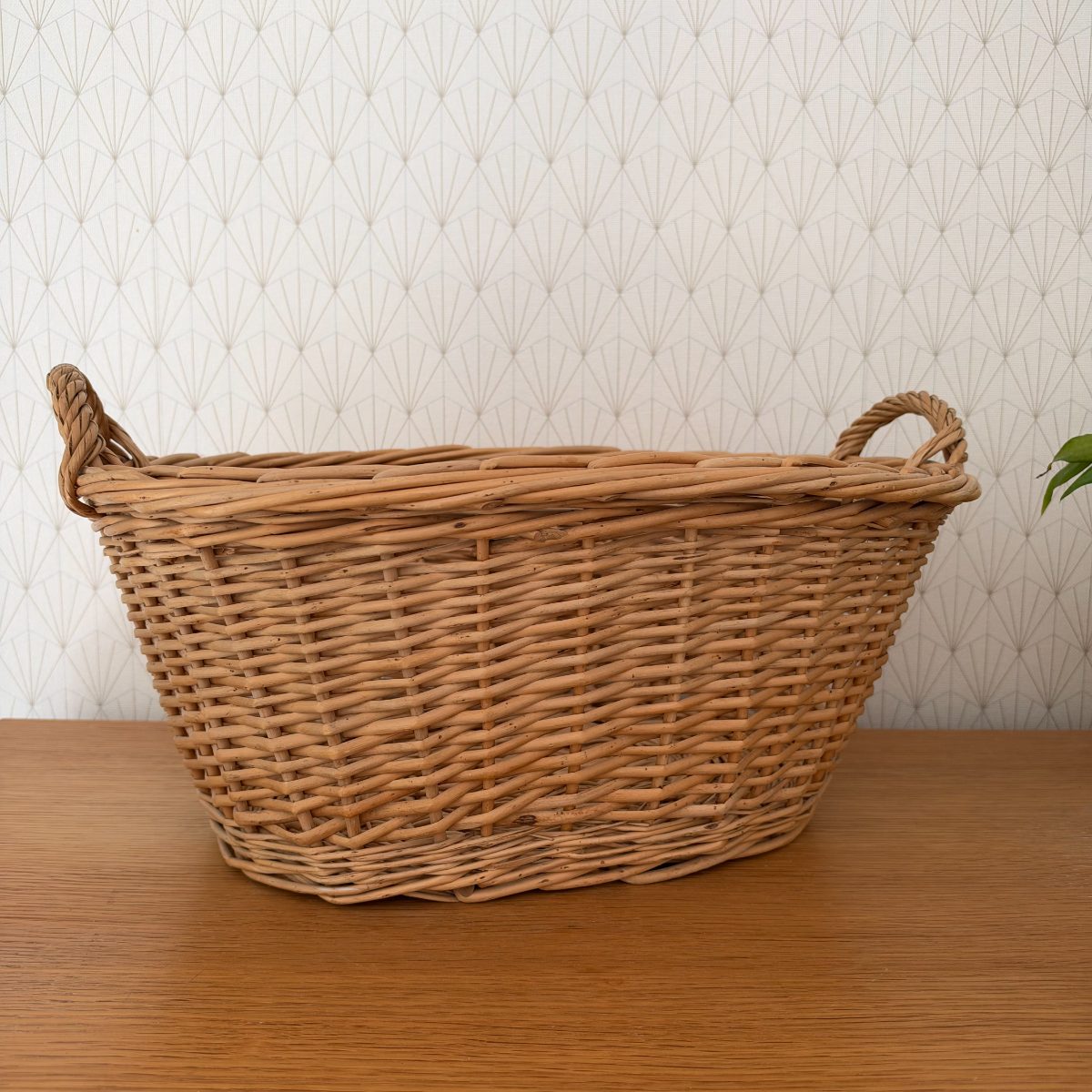 Vintage French wicker laundry basket with two loop handles, oval shape, natural honey tone, photographed from the front on a wood surface.