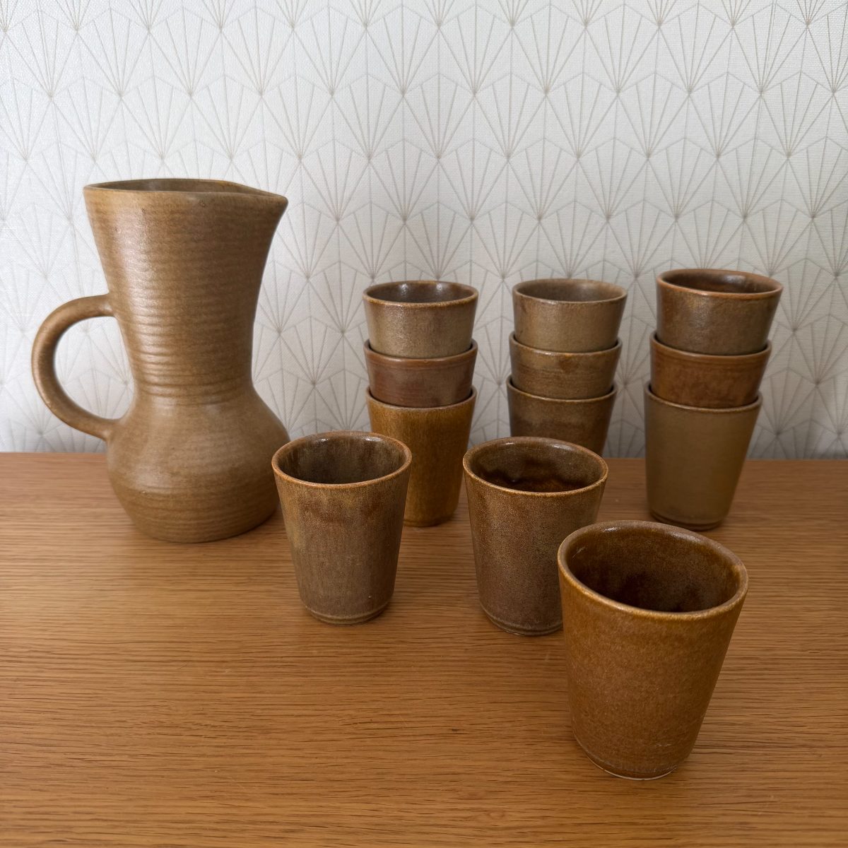 Full view of French midcentury stoneware pitcher with ribbed body and handle alongside twelve small stoneware cups in warm brown tones
